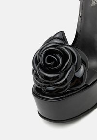 Chaussure à talon haut noire avec une surface brillante. Présente un détail en forme de rose noire proéminent à l'avant et une semelle épaisse.