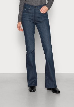 Osoba nosząca ciemnoniebieskie jeansy typu bootcut, szary prążkowany sweter, czarne buty na obcasie, stojąca na jednolitym jasnym tle.