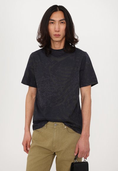 Tricou cu mânecă scurtă de culoare bleumarin, cu un model abstract în nuanțe închise, asortat cu o pereche de pantaloni texturați kaki. Modelul ține o geantă mică de culoare neagră.