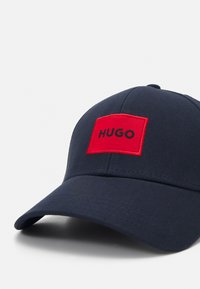 Mornariće plava kapa od pamuka sa strukturiranim oblikom. Ima crveni pravokutni natpis s "HUGO" u crnom slovu.