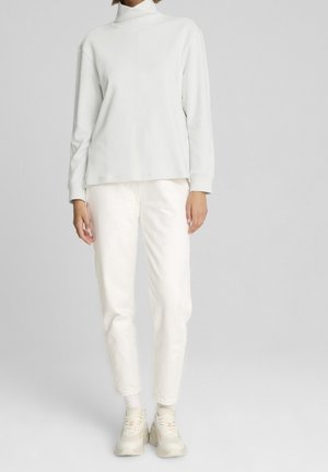 Osoba oblečená v bílém roláku, bílých teplákových kalhotách, bílých ponožkách a robustních světle béžových teniskách, stojící na šedém pozadí.