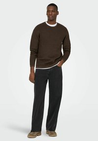 Pull en brun texturé avec une couche intérieure blanche, associé à un jean large gris foncé et des chaussures beiges. Un modèle masculin debout pose de manière décontractée.