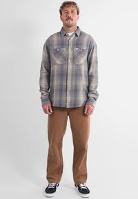 Chemise en flanelle à carreaux gris et beige avec deux poches poitrine, à boutons, manches longues, associée à un pantalon beige et des baskets noires.
