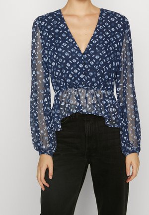 Frau in einer dunkelblauen, geknöpften Bluse mit weißem geometrischen Muster, durchsichtigen langen Ärmeln und schwarzen hochgeschnittenen Hosen, die vor einem einfarbigen Hintergrund steht.