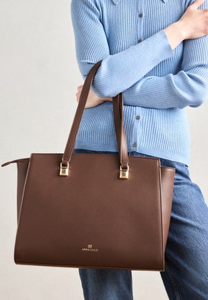 Brun læder tote bag med hardware i guld-tone, struktureret form og præget logo. Har dobbelte håndtag og en glat tekstur.