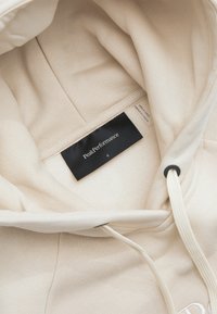 Beige Peak Performance hoodie met zichtbaar zwart merklabel en maat "S" aan de binnenkant van de capuchon, crèmekleurige trekkoorden met zwarte oogjes.