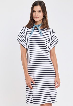 Femme portant une robe blanche avec des rayures bleu marine horizontales et verticales et un foulard bleu clair autour du cou, debout devant un fond uni.