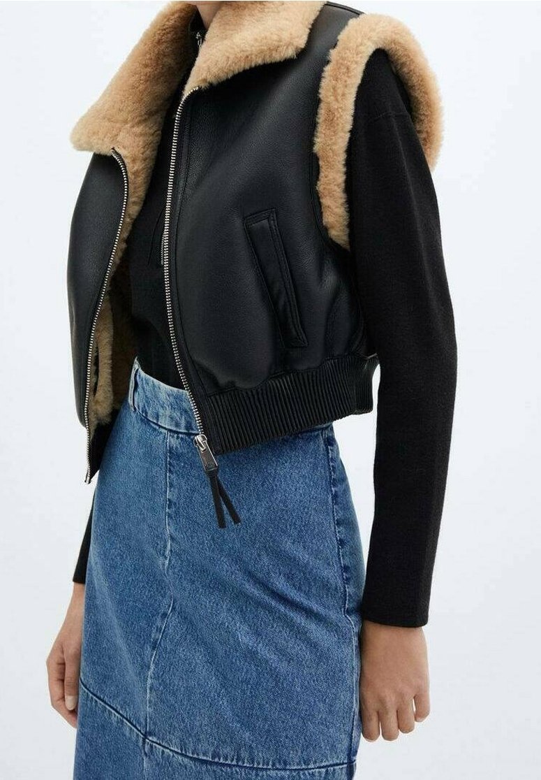 Veste en cuir noire sans manches avec col en shearling beige, portée sur un haut à manches longues noir, associée à une jupe en denim bleu taille haute.