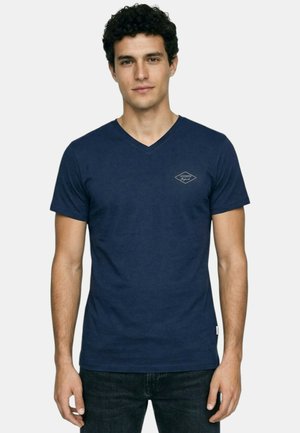 Jeune homme aux cheveux bouclés et foncés portant un T-shirt bleu marine à encolure en V avec un petit logo sur le côté gauche de la poitrine, debout devant un fond uni.