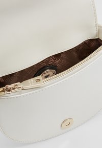 Bílá taška z umělé kůže se zlatým zipem a kováním. Uvnitř je viditelná hnědá vzorovaná podšívka s detailem logo disku.