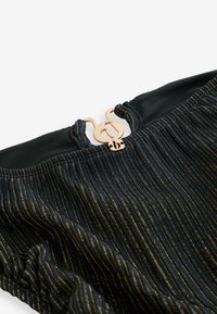 Tissu noir avec des rayures verticales dorées, encolure volantée, et fermeture à double anneau métallique doré avec un petit charm en forme de "b".