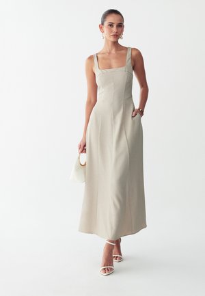 Vrouw draagt een beige mouwloze maxi-jurk, houdt een kleine witte handtas vast, geaccessoriseerd met pareloorbellen en witte open-toe hakken.