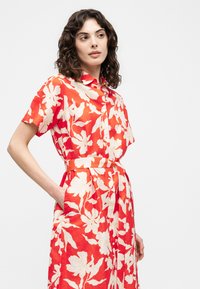 Γυναίκα με σγουρά μαλλιά μήκους μέχρι τους ώμους, φοράει κόκκινο φόρεμα πουκαμίσα με λευκά λουλουδένια σχέδια και φαρδύ ζωνάκι στο ίδιο χρώμα, έχει το χέρι στην τσέπη και κοιτάζει πλαγίως.