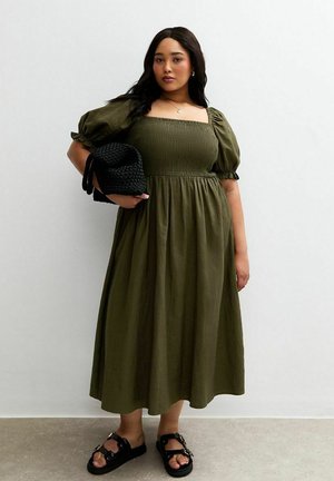 Olivgrünes Kleid mit Puffärmeln, quadratischem Ausschnitt und gerafftem Oberteil, das bis zu den Knöcheln fällt. Getragen mit schwarzen Sandalen und einer geflochtenen Tasche.