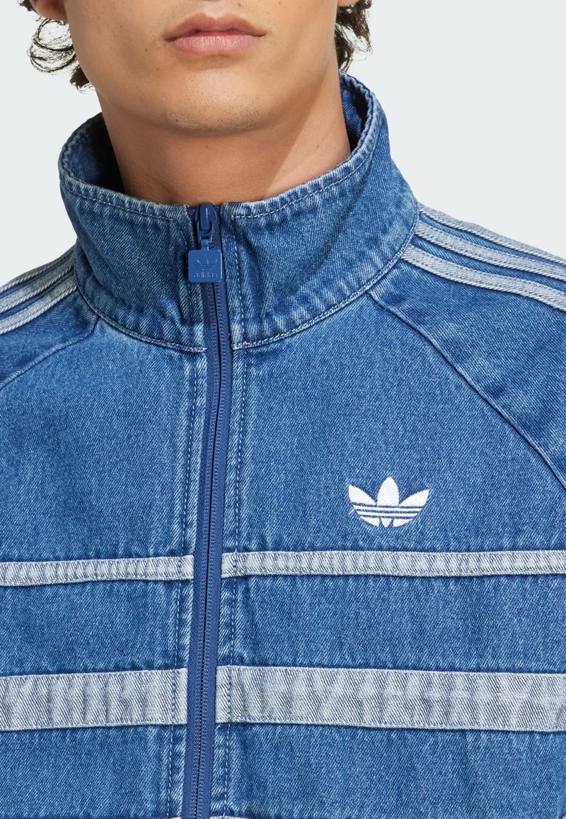 Lysblå denimjakke med opretstående krave, frontzipper og hvide vandrette striber. Har broderet logo på venstre bryst.