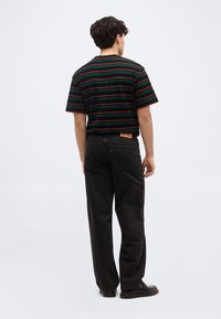 Άνδρας με κοντά σγουρά μαλλιά, ντυμένος με μαύρο και πράσινο ριγέ μπλουζάκι, μαύρο τζιν και μαύρα παπούτσια, στέκεται με την πλάτη στην κάμερα.