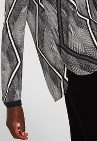 Blusa a maniche lunghe con motivo geometrico in bianco e nero indossata con pantaloni neri, con un braccio rilassato e la mano appoggiata di lato.