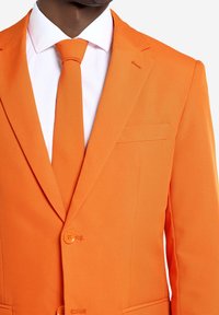 Oranžs uzvalka žakets ar ielaiduma atlokiem, diviem pogām un krūšu kabatu. Kombinēts ar baltu kleita kreklu un atbilstošu oranžu kaklasaiti.