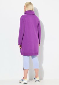 Langer lila Sweatshirt mit hohem Kragen, gerippten Bündchen und Saum. Kombiniert mit hellblauen Leggings und mehrfarbigen Turnschuhen.