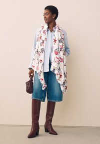 Sjaal met bloemenprint, lichtgewicht stof, gedrapeerd over een blauw gestreept overhemd, gecombineerd met denim shorts en bruine, kniehoge laarzen met hak.