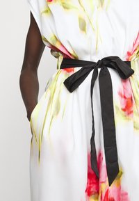 Blommönstrad klänning i vitt, med abstrakta röda och gula mönster, samt ett svart tygbälte knutet i midjan och sidofickor.