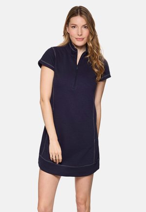 Mujer con cabello largo y ondulado, vestida con un vestido corto azul marino con cremallera de manga corta, de pie frente a un fondo claro y sencillo.