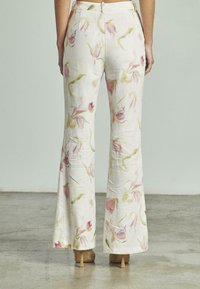 Vrouw in witte wijde broek met roze en groene bloemenprint, staande op een grijze betonnen vloer, getoond vanuit de achterkant.