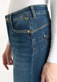 Jeans in denim blu aderenti con cuciture dorate, dotati di vita media, tasche frontali e accessori metallici. Texture liscia con lieve scolorimento.