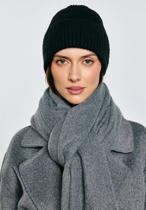 Černá pletená čepice, šedý vlněný šál a kabát. Šál je silný a volně se omotává kolem krku. Dominují měkké textury a neutrální barvy.