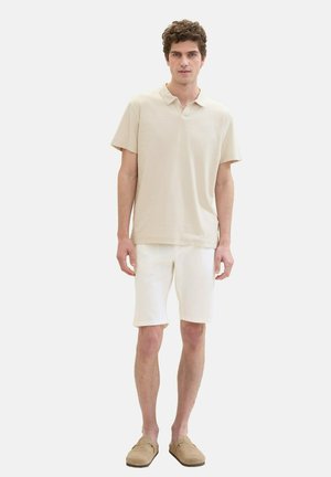 Beige short-sleeve polo shirt with a collared neck, paired with white shorts. The model wears light tan slip-on shoes, standing against a white background.