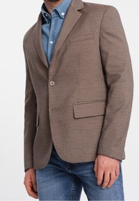 Brązowo-beżowa marynarka w pepitkę z wyciętymi klapami, dwoma przednimi kieszeniami i pojedynczym guzikiem, noszona na koszuli dżinsowej.