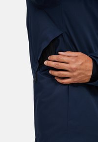 Námořnická bunda s hladkým, matným povrchem, která má boční kapsu na zip a detailní záběr ruky, jak pevně uchopuje otvor kapsy.