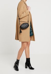 Bolso cruzado acolchado negro con forma redondeada, detalle de borlas y correa ajustable. Combinado con un abrigo beige y una falda de cuadros.