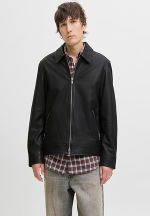 Jeune homme portant une veste en cuir noire zippée par-dessus une chemise à carreaux rouge et un jean délavé, debout devant un fond clair uni.