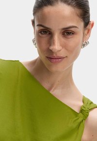 Top vert à une épaule avec un détail de sangle nouée. Tissu lisse et léger avec une texture subtile. Mis en valeur par des boucles d'oreilles en forme de cercle argentées.