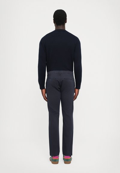 Homme se tenant de dos, portant une chemise à manches longues marine foncé, un pantalon assorti et des chaussures vertes avec des chaussettes roses sur un fond blanc uni.