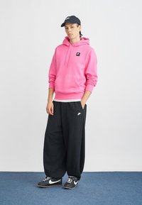 Sweatshirt cor-de-rosa com capuz, bolso frontal e logótipo preto, combinado com calças de suor pretas de perna larga com logótipo branco, e ténis pretos.