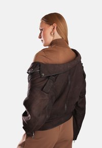 Hnědá kožená bunda s detaily na zip, volný střih a oversize límec. Nositá na ramenou, ukazující hnědý rolák pod ní.