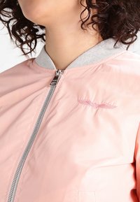 Bomber jacket rosa chiaro con colletto grigio, zip argentata e logo ricamato. Texture liscia con design aderente, dotata di tasche laterali.