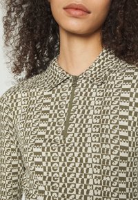 Ziptröja i olivgrön färg med upprepat mönster av text i off-white. Bomullsblandad tyg med krage och korta ärmar.