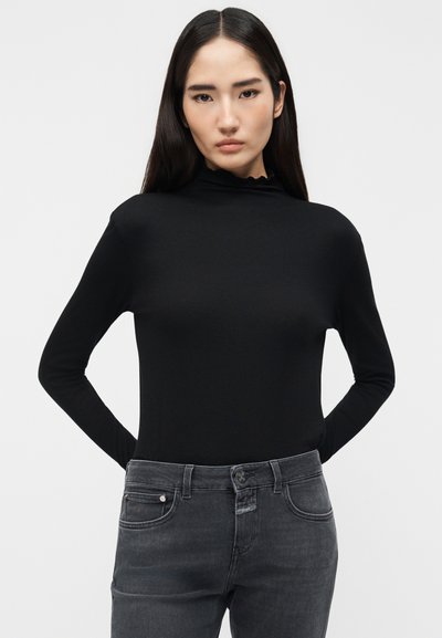 Femme aux longs cheveux foncés portant un haut noir à manches longues et un jean gris foncé devant un fond clair uni.