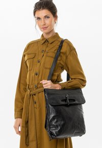 Borsa in pelle nera con chiusura a pattina e dettagli hardware sottili, portata da una persona che indossa un vestito giallo senape con bottoni.