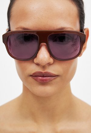 Brune oversized solbriller med lilla tonede linser, der har en tyk, rektangulær ramme. Glat overflade uden synlige mønstre eller udsmykninger.