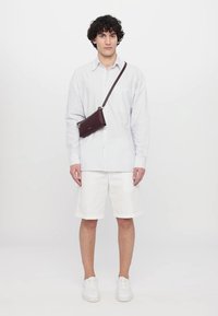 Brun crossbody-veske med stropp, båret over en lyseblå stripete skjorte og hvite shorts. Utstyrt med hvite sneakers og en nøytral bakgrunn.