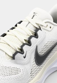 Bijela atletska cipela s prozračnom mrežastom gornjištu, kontrastnim sivim detaljima i gumenom potplatom. Ima ravne vezice i Nike logo.