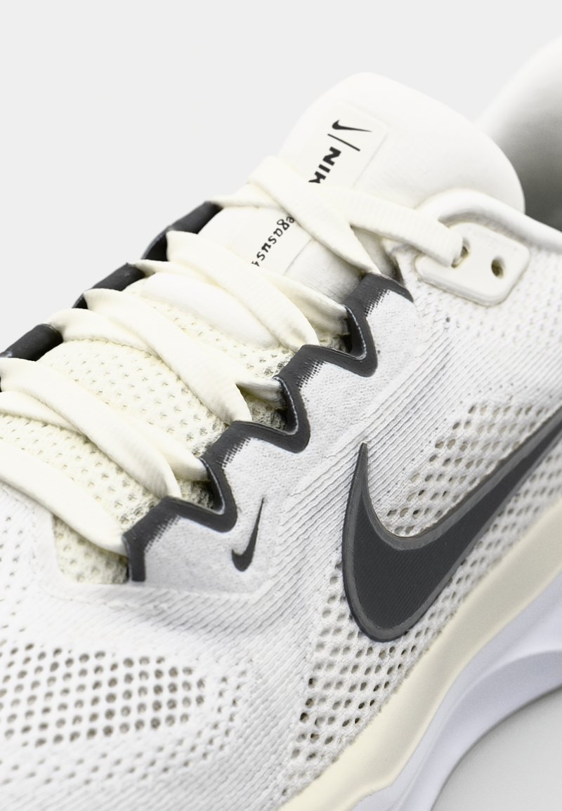 Zapatilla deportiva blanca con parte superior de malla transpirable, acentos grises contrastantes y suela de goma. Cuenta con cordones planos y un logo de Nike.