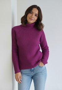 Fioletowy prążkowany sweter z golfem i długimi rękawami, o luźnym kroju i teksturowanym splocie. W połączeniu z jasnoniebieskimi dżinsami.