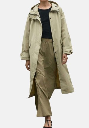 Trench coat impermeable en color beige con capucha, cierres a presión y bolsillos laterales. Combinado con pantalones holgados en color caqui y sandalias negras.