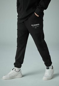 Svarta sweatpants med texturerade paneler, prydda med en vit "ALLSAINTS" logotyp. I kombination med kraftiga vita sneakers och elastiska muddar.