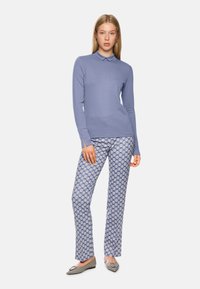 Langärmliges, hellblaues Poloshirt kombiniert mit gemusterten blauen Hosen mit weißen Motiven; das Modell trägt graue flache Schuhe.